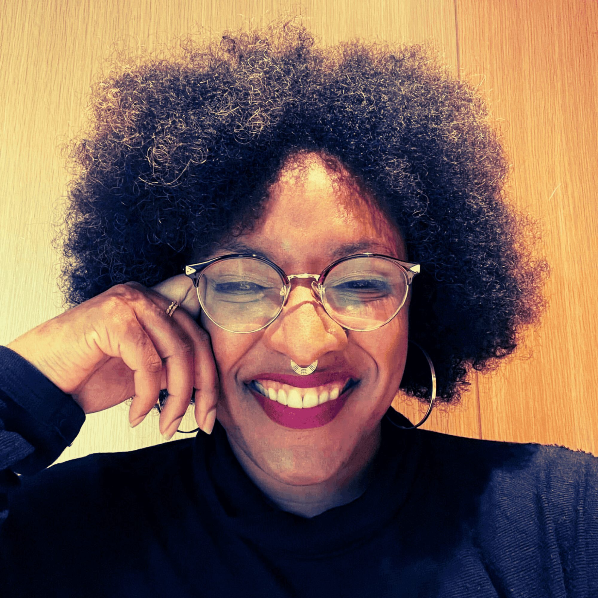 Smiling woman with a large afro, round glasses, and septum piercing, leaning on her hand.